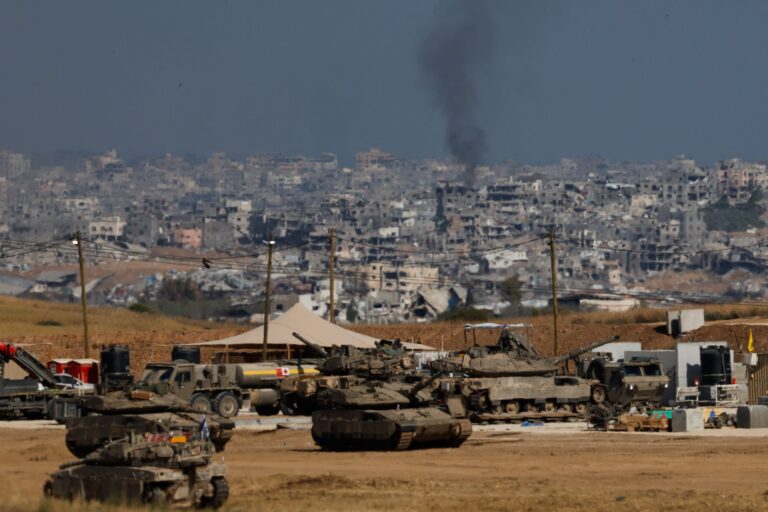 Military vehicles, including tanks and armoured trucks, are positioned near the border. In the background, smoke rises over a densely built urban area with damaged buildings.