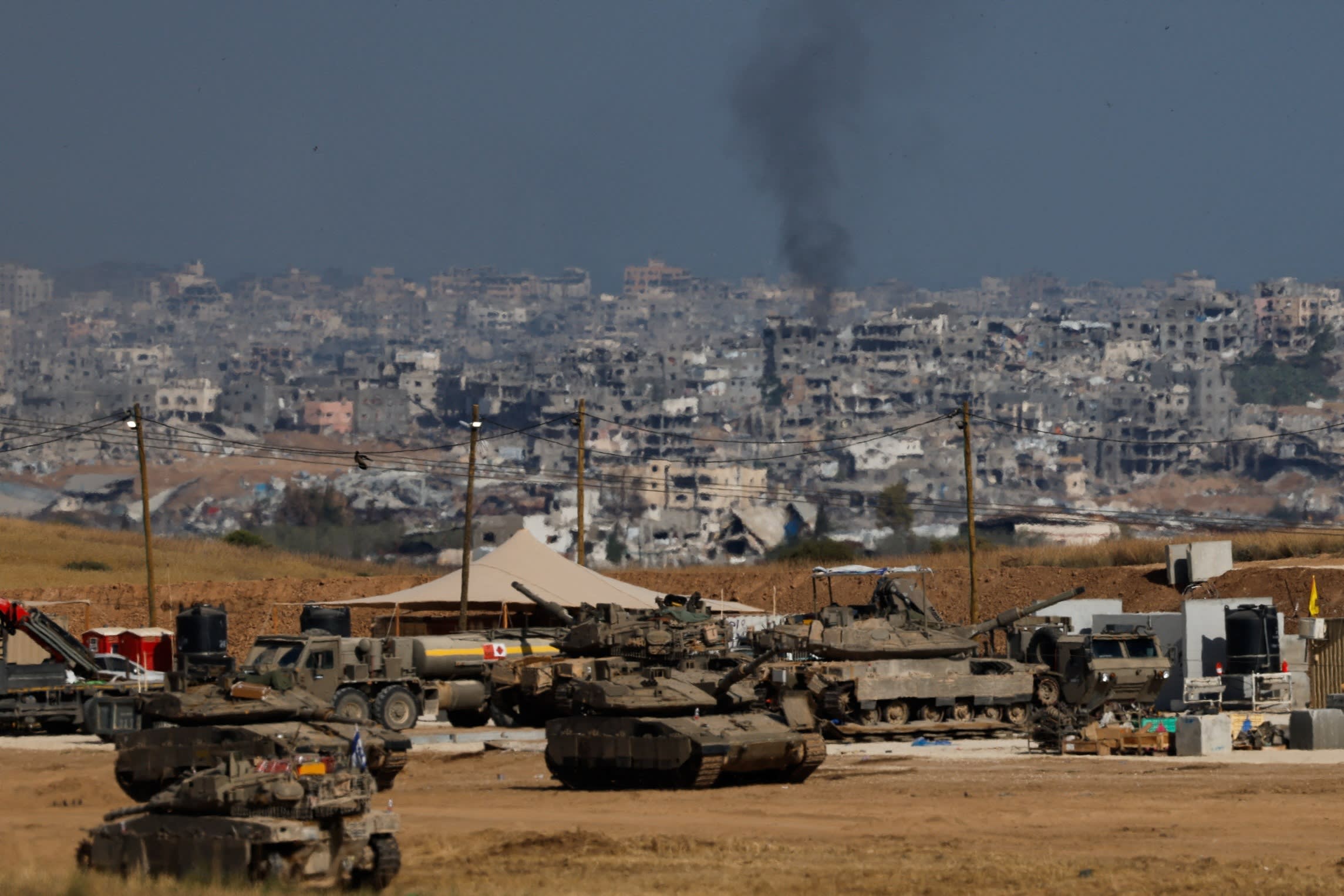 Military vehicles, including tanks and armoured trucks, are positioned near the border. In the background, smoke rises over a densely built urban area with damaged buildings.