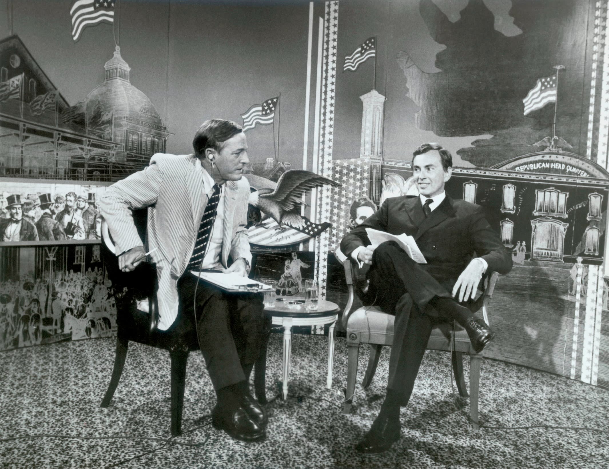 Two men in suits and ties, seated in armchairs in a room or studio that is carpeted and decorated with US flags and scenes from US history