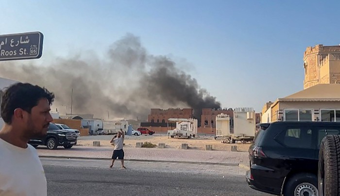 Smoke rises from buildings in Doha after explosions, with people standing on the street in the foreground.