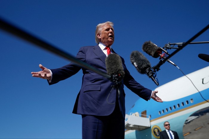 Donald Trump speaks to reporters in front of microphones with Air Force One visible behind him.