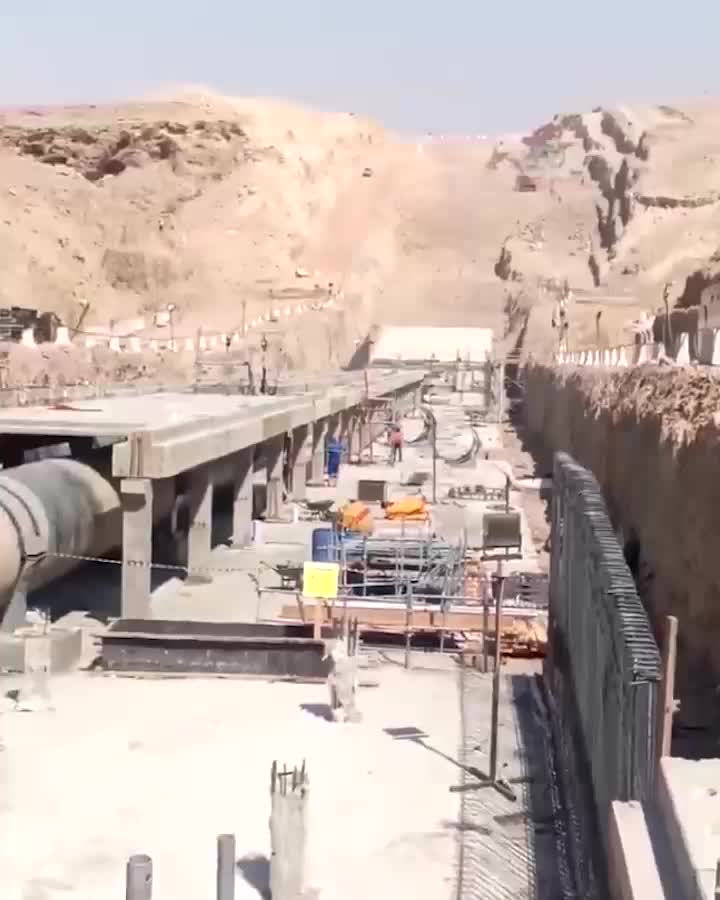 Concrete infrastructure under construction in a dry, rocky cut, showing a long trench, scaffolding, large pipes and a few workers.
