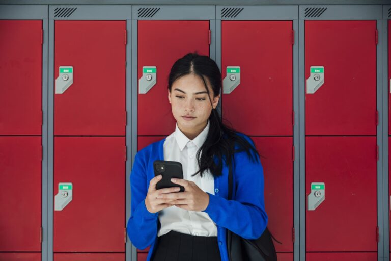 A medium close-up front view of a teenage girl who is sending text messages to a friend and checking her social media as she waits in the corridor of the high school she attends. 