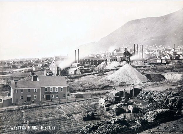 Historical photograph showing C&C Shaft mining complex with industrial buildings, smokestacks, tailings piles, and Virginia City residential buildings against mountainous Nevada terrain