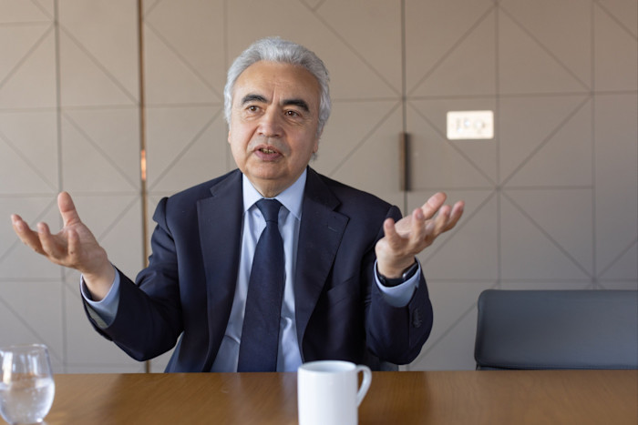 Fatih Birol gestures with both hands while seated at a table with a glass of water and a white mug.