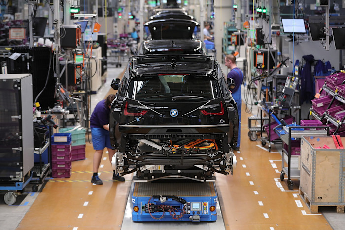 BMW i3 electric cars move down an assembly line as employees work on them at a factory, surrounded by tools and equipment.